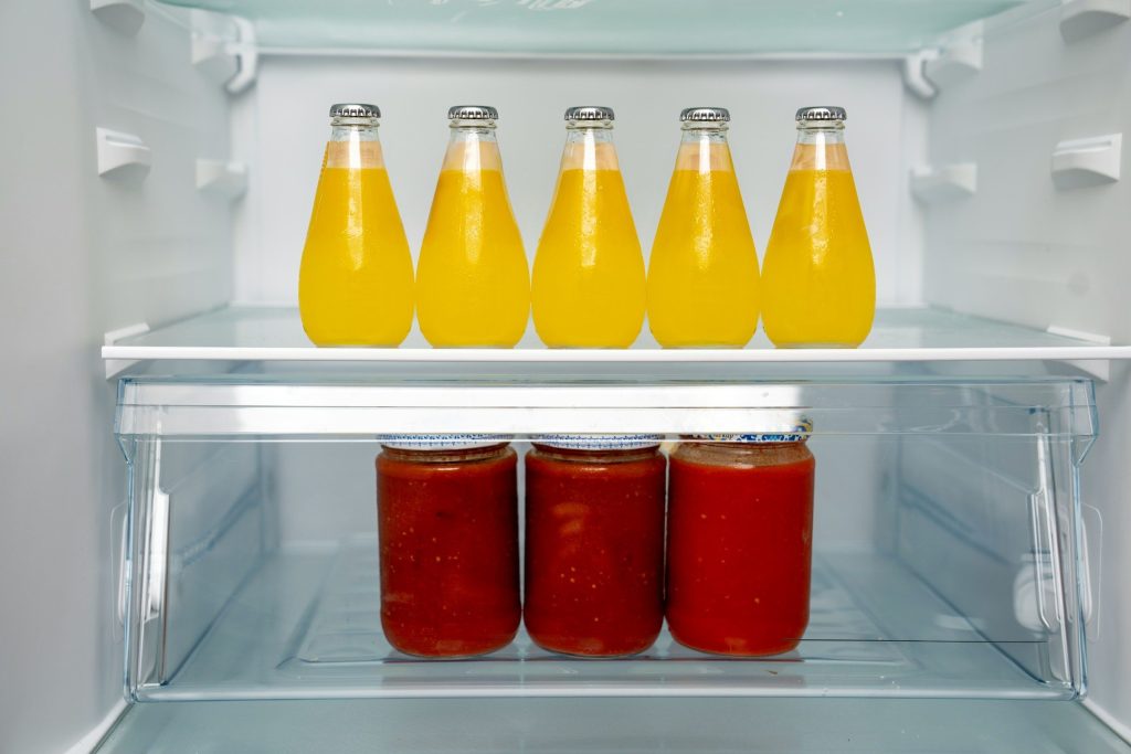 Five bottles of juice on a glass shelf above three jars of sauce.
