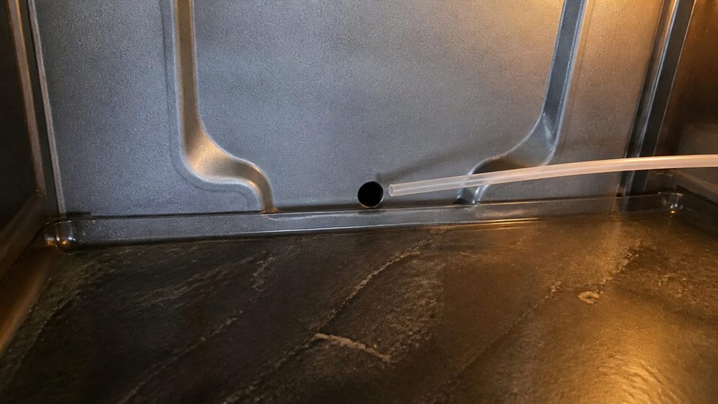 A close view of the small defrost drain hole at the base of the rear wall inside a refrigerator compartment, the shallow channel around it visible, a thin flexible tube approaching it.