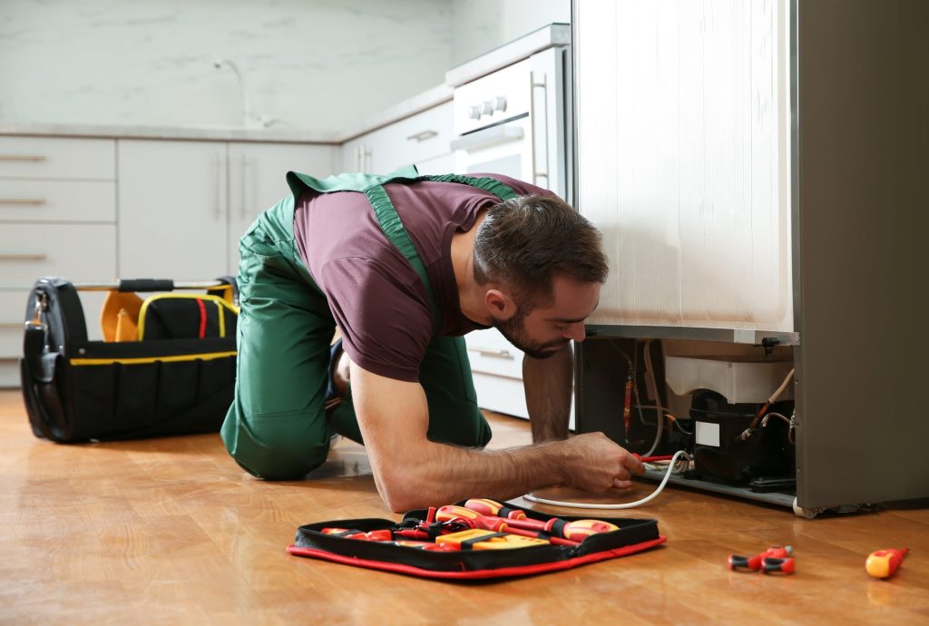 A technician working behind a refrigerator, tool roll on the floor.