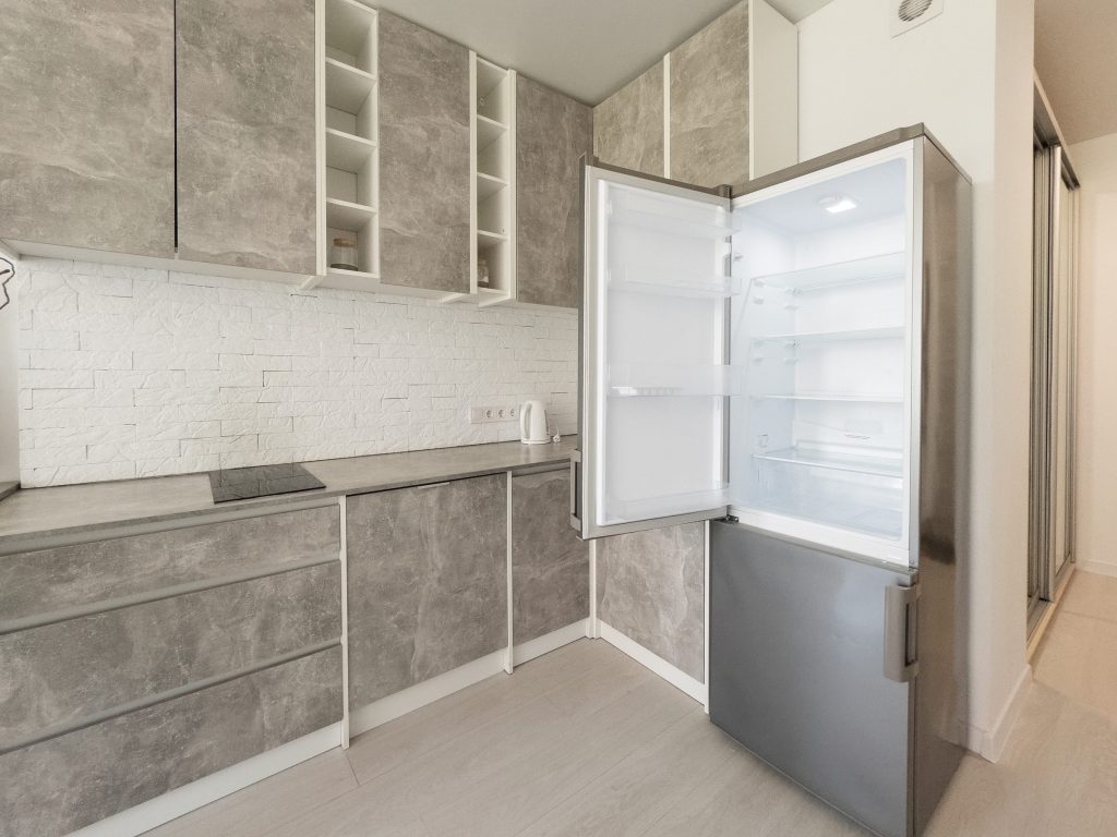 A compact bottom-freezer refrigerator standing open and empty in a pale tiled kitchen, the freezer door below ajar