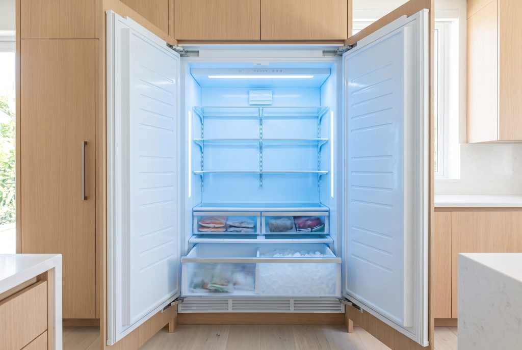 An integrated freezer column standing open, interior drawers pulled out on frosted packets and an ice bin, cold blue light against a pale oak kitchen