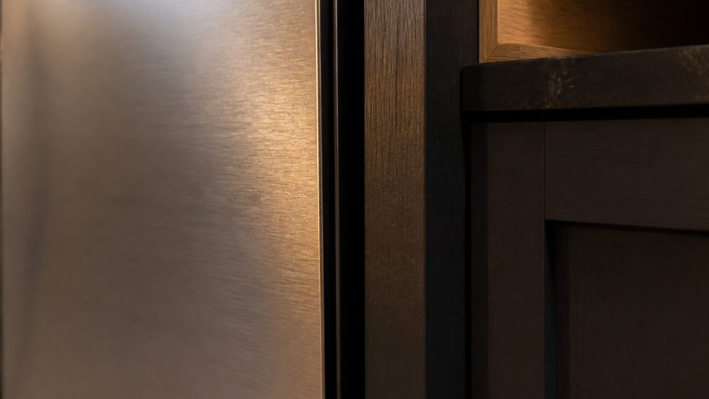 The narrow shadow gap between a refrigerator's side and the cabinet carcass beside it, brushed metal against dark timber, raking amber light.