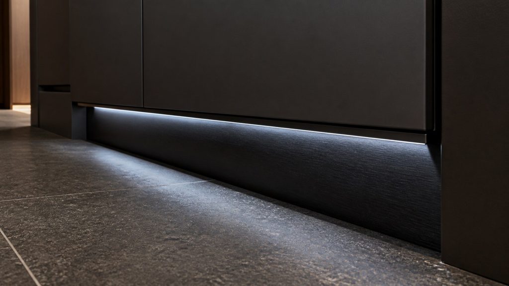 A close view along the toe kick under a tall refrigerator, dark brushed panel meeting dark stone floor, a thin line of cold light above.
