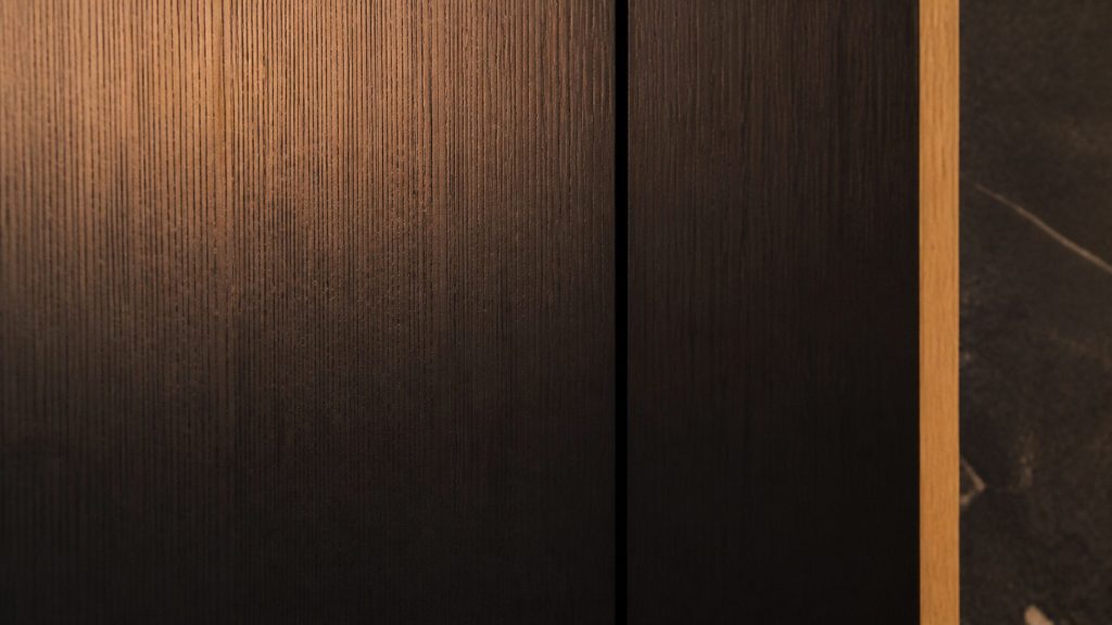 The joint between an integrated refrigerator's cabinet front and the panel next to it, a fine shadow line down dark timber, amber light across the grain.
