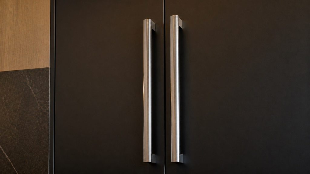 Two long vertical handles side by side on adjoining refrigeration columns, brushed steel against near-black fronts, close and slightly from below.