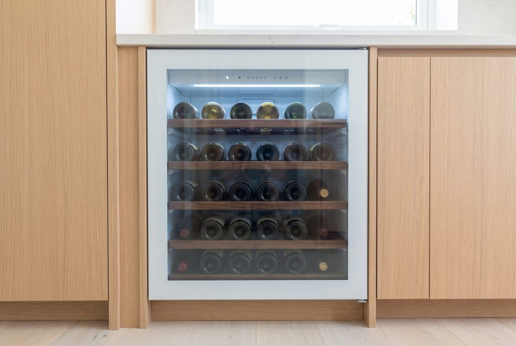 An undercounter wine cabinet with a glass door set into a run of oak cabinetry, bottles lit on wooden shelves inside