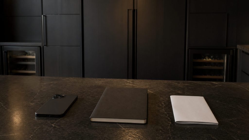 Three things laid in a row on a dark stone worktop under warm light: a phone face down, a closed notebook, and a single folded sheet of paper.