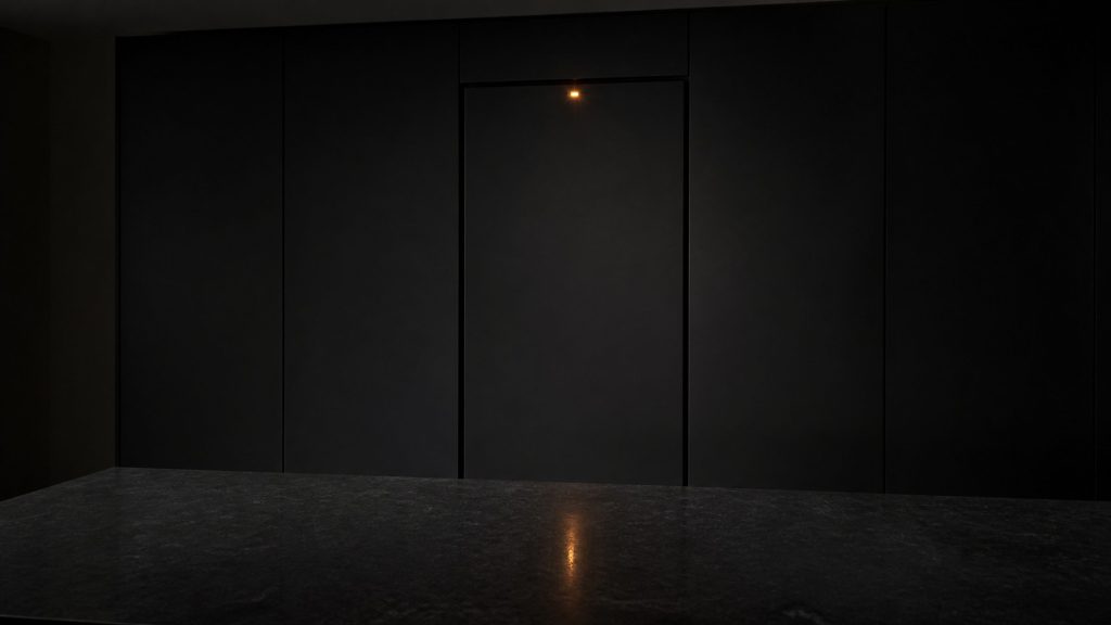 A single small amber warning light glowing on the narrow top edge of a tall integrated refrigerator door in an unlit kitchen at night, the rest of the room in near-black, one amber reflection on the dark stone worktop.